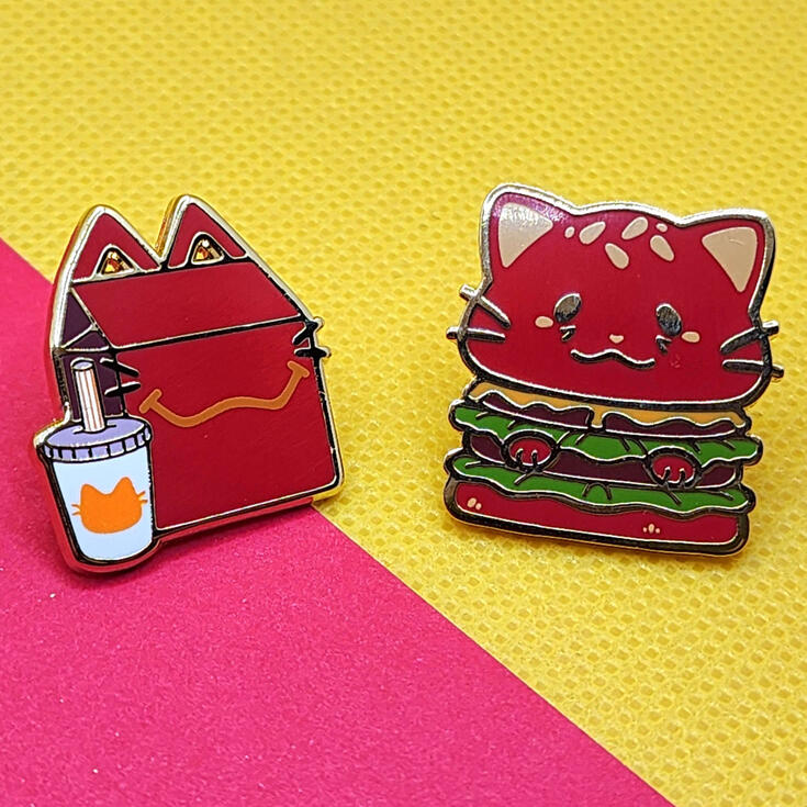 Catfood Enamel Pins photograph of two enamel pins, one pin designed to look like a cat happy mean (red box, white soda cup, yellow cat mouth and cat head printed) and the other like a cat burger (red cat shaped bun, regular burger interior)