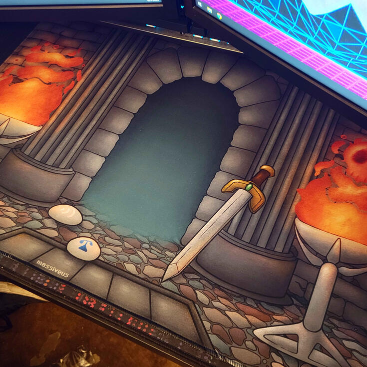 OSRS Desk Mat photograph of a desk mat sitting under a computer monitor. the deskmat depicts the log in screen from OSRS which is a dungeon style doorway with columns on either side and fire braziers that contain fire shaped like runes