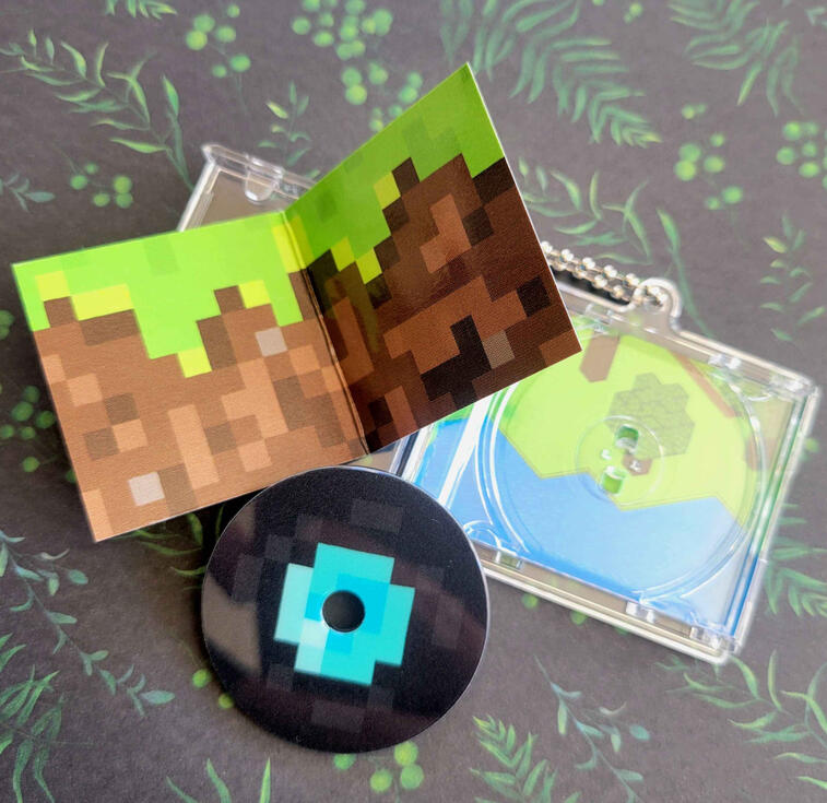 Minecraft CD Keychain photograph of a CD style keychain. the photo shows the interior cover booklet that looks like a dirt block from minecraft, the removable acrylic CD that looks like the record item from minecraft, and the crystal plastic CD casing that has more minecraft de